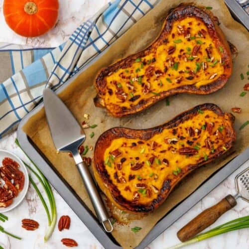 Two roasted butternut squash halves filled with creamy Parmesan mash, topped with pecans and scallions, on a parchment-lined baking sheet.