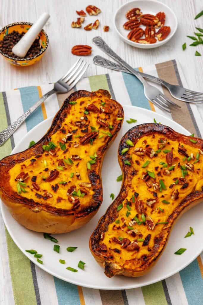 Two stuffed and broiled butternut squash halves on a white plate, topped with pecans and scallions, surrounded by forks and small bowls of ingredients.