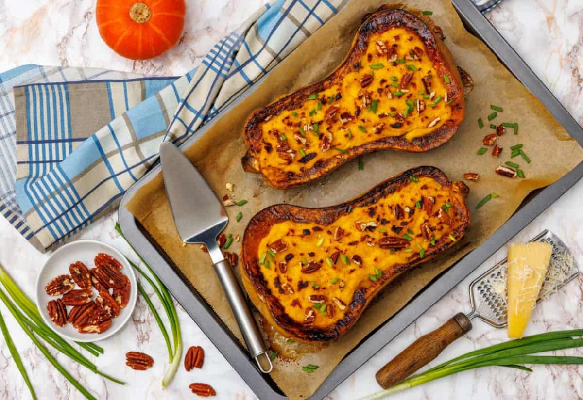 Two roasted butternut squash halves filled with creamy Parmesan mash, topped with pecans and scallions, on a parchment-lined baking sheet.