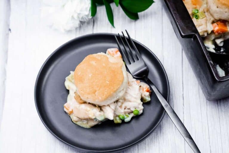 A black plate features a biscuit-topped serving of chicken pot pie, complete with chicken, peas, and carrots. A fork is placed on the plate, with a baking dish partially visible in the background.