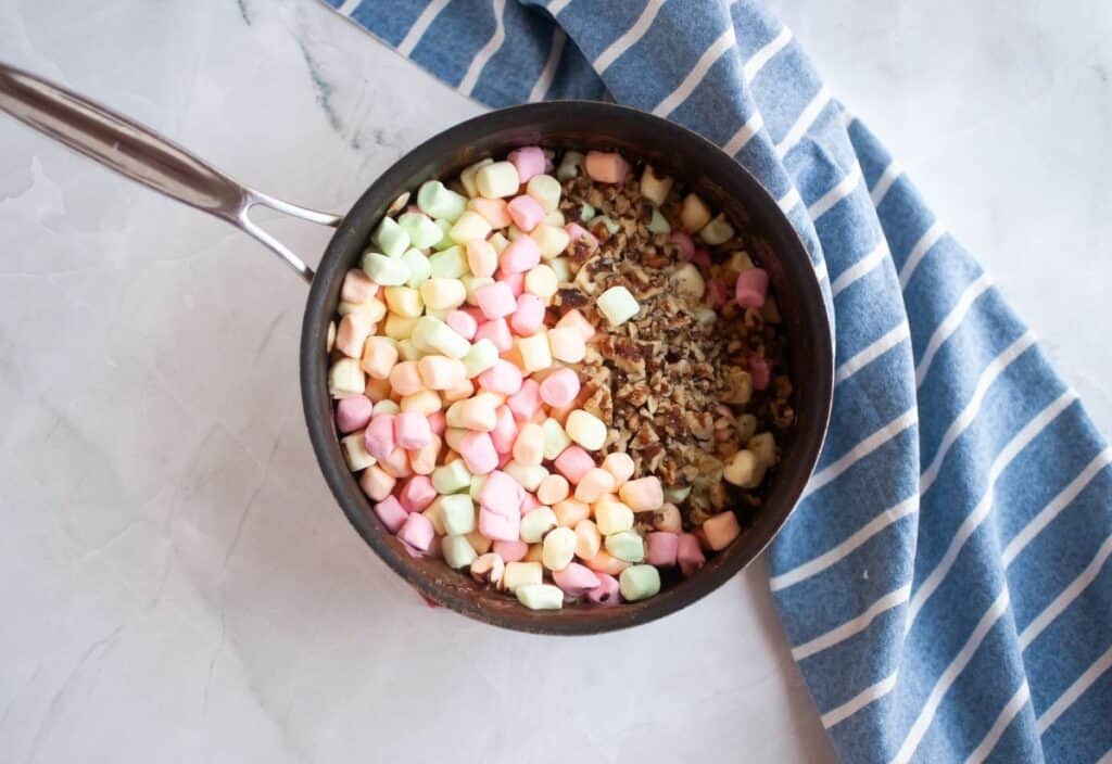 Pastel marshmallows and chopped walnuts added to the melted chocolate mixture in a saucepan.