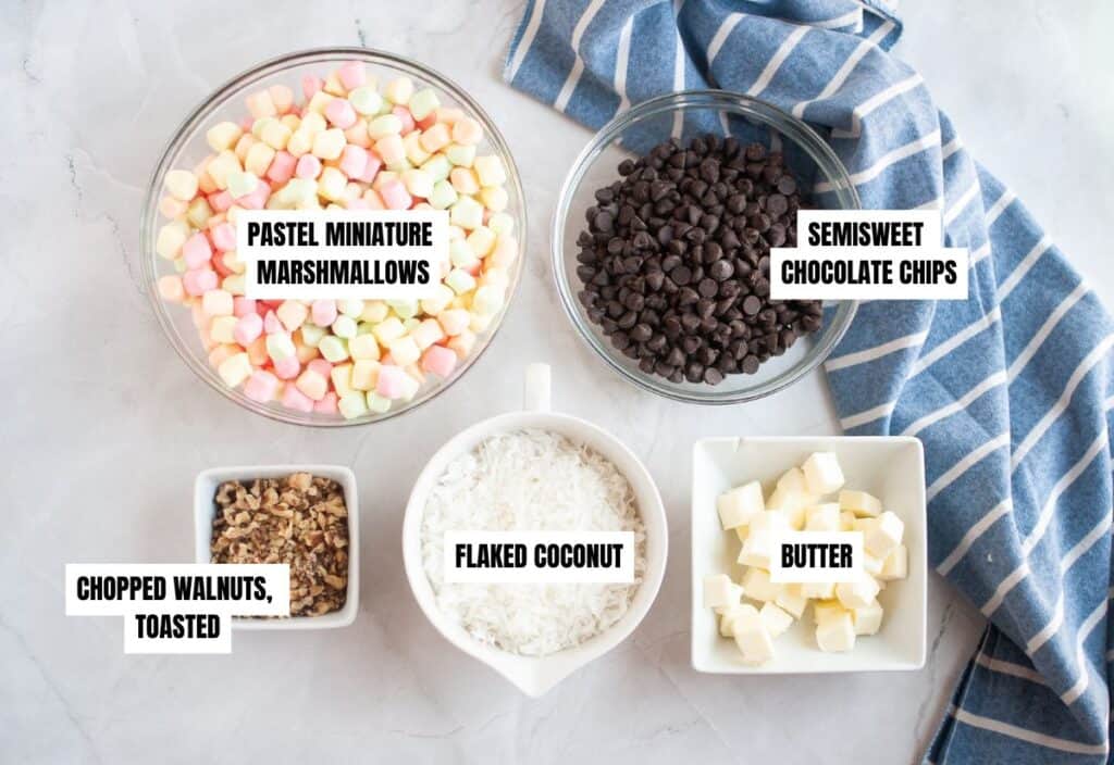 A flat-lay photo of all the recipe ingredients arranged neatly: bowls of pastel miniature marshmallows, semisweet chocolate chips, flaked coconut, chopped toasted walnuts, and cubed butter, with a blue striped towel beside them.