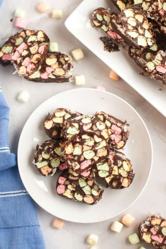 A top-down shot of several slices of church window cookies arranged on a white plate and platter, with scattered mini marshmallows around the scene and a blue cloth nearby.