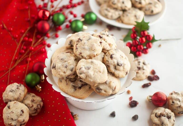 A white pedestal plate piled with cranberry chocolate chip cookies surrounded by red and green Christmas ornaments and berries on a red cloth.