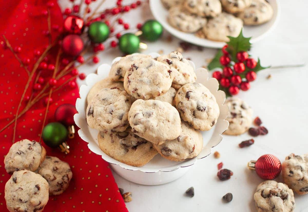 A white pedestal plate piled with cranberry chocolate chip cookies surrounded by red and green Christmas ornaments and berries on a red cloth.