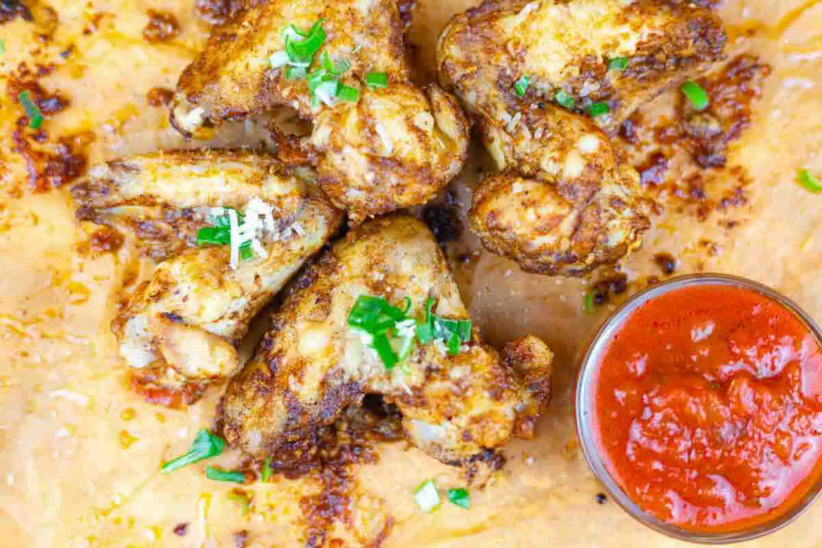 Five seasoned chicken wings on parchment paper, garnished with chopped herbs, next to a small bowl of red dipping sauce.