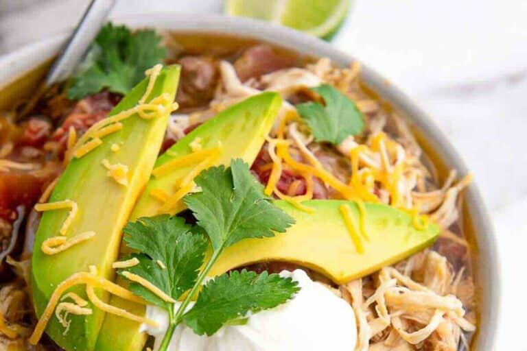 Chicken tortilla soup topped with avocado slices, shredded cheese, sour cream, and cilantro, with lime wedge nearby.