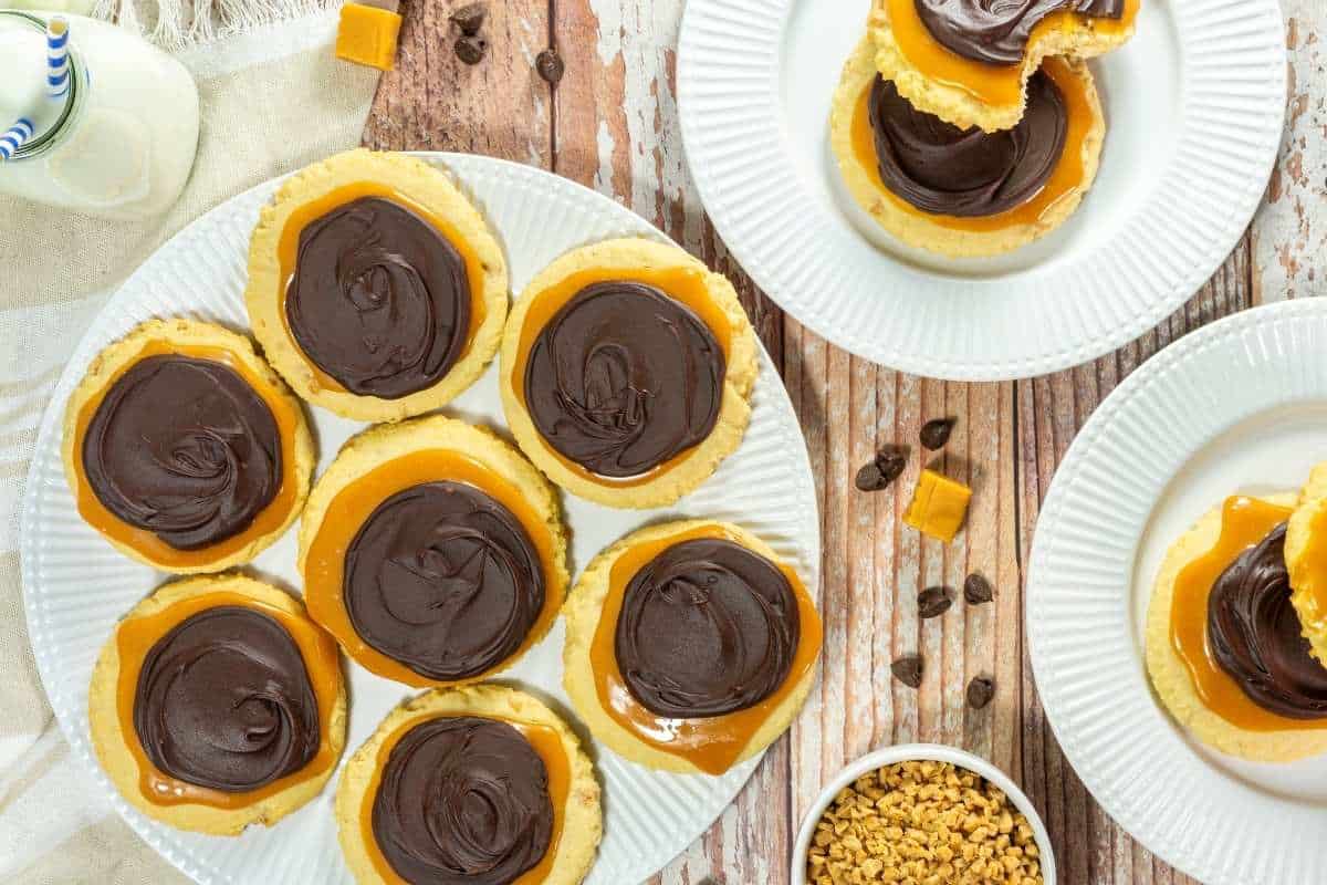 A plate of Copycat Crumbl Twix Cookies, with more cookies on separate plates, caramel pieces, and small chocolate chips nearby on a wooden table.