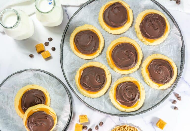 Overhead view of Crumbl copycat Twix cookies on trays and plates, topped with caramel and chocolate, with milk bottles, caramels, and chocolate chips around them.
