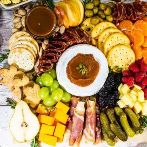 Overhead view of a colorful fall charcuterie board with brie, crackers, cheeses, cured meats, fruits, pickles, and autumn-themed cookies arranged on a wooden surface.