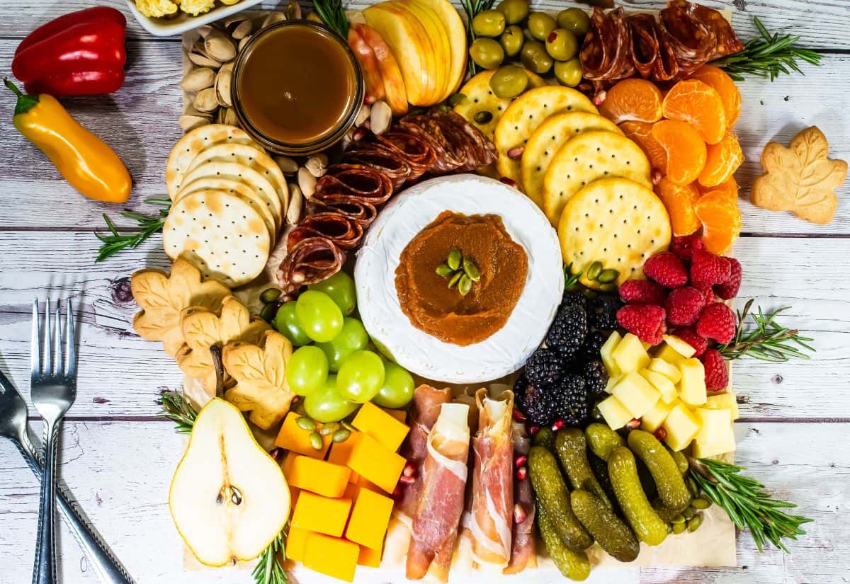 Overhead view of a colorful fall charcuterie board with brie, crackers, cheeses, cured meats, fruits, pickles, and autumn-themed cookies arranged on a wooden surface.
