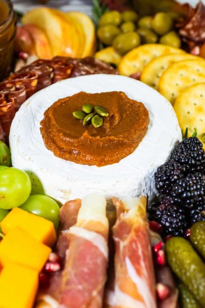 Brie cheese topped with pumpkin butter and pepitas at the center of a charcuterie board, surrounded by grapes, blackberries, cured meats, and crackers.