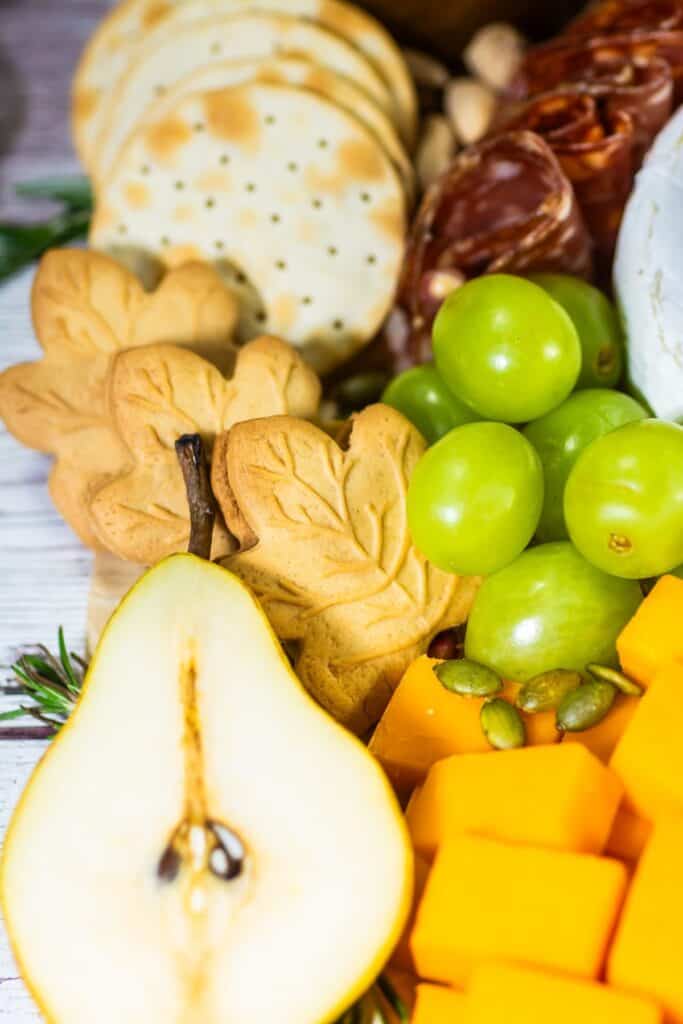 Close-up of a fall charcuterie board with sliced pear, cheddar cubes, green grapes, leaf-shaped cookies, and round crackers.