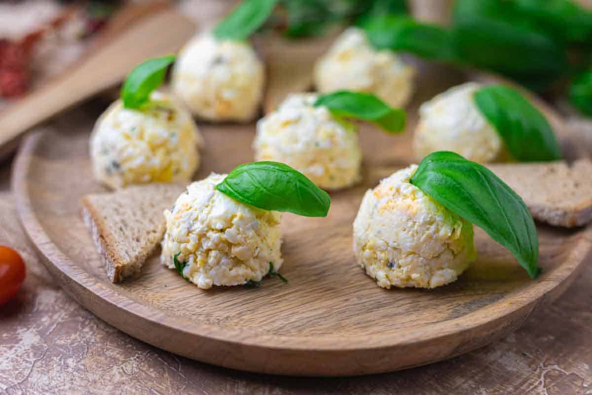 Cheese balls garnished with fresh basil leaves on a wooden plate, accompanied by triangular bread slices.