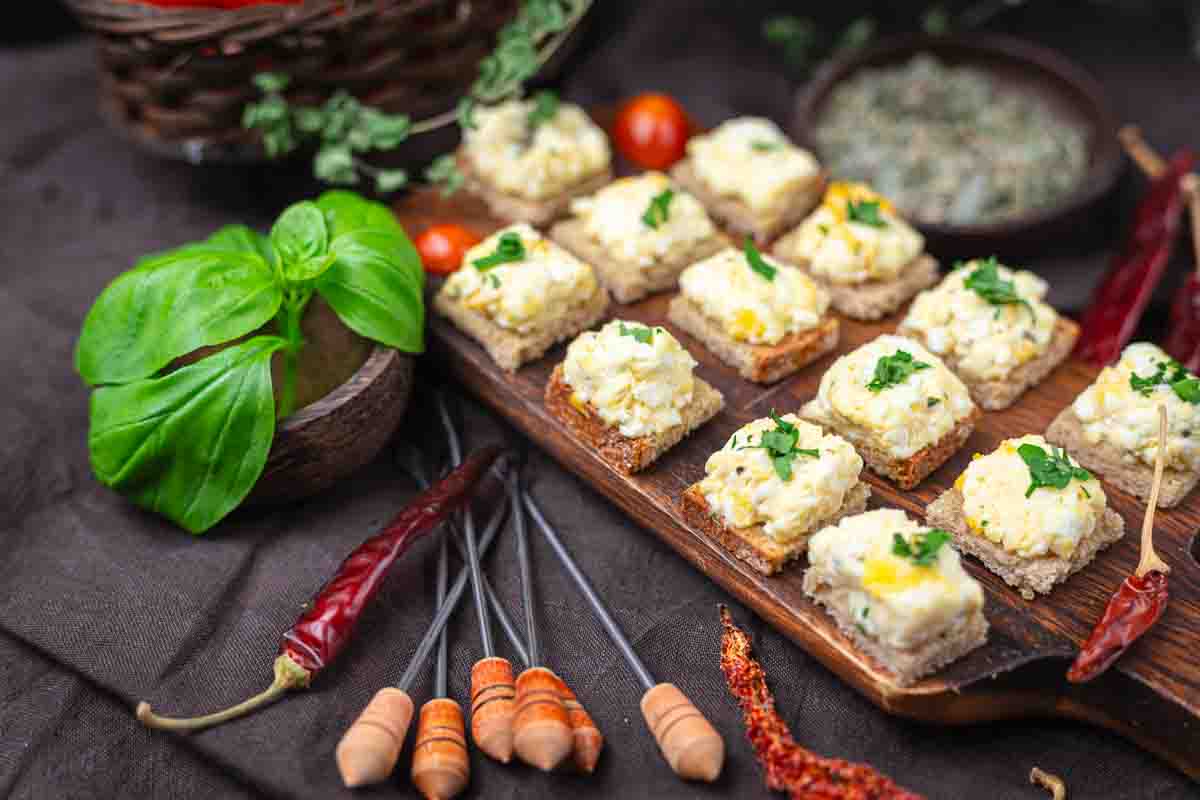 Several pieces of bread topped with a creamy feta and herb eggs, arranged on a wooden board, surrounded by fresh basil, dried chilies, and cherry tomatoes.