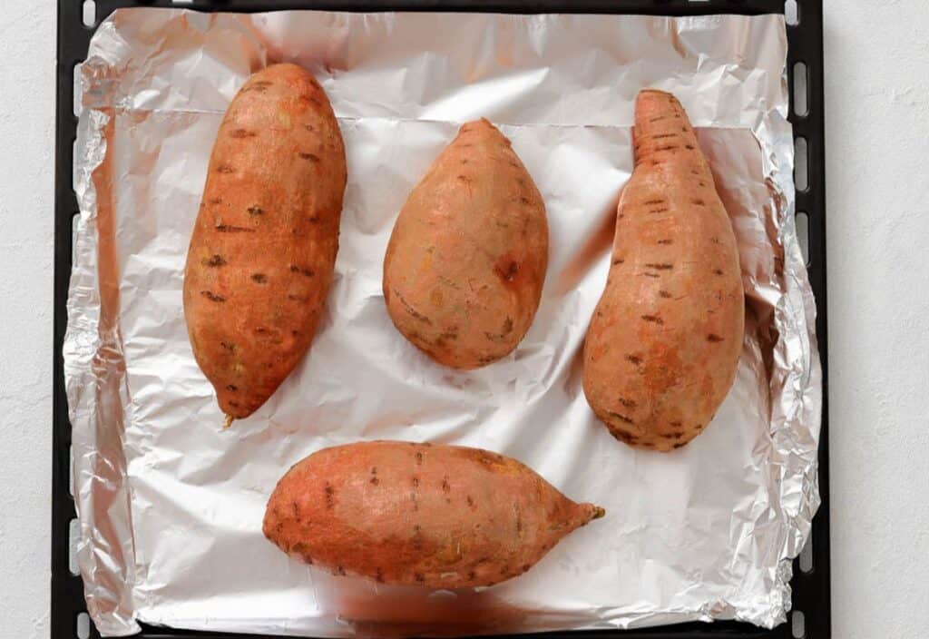Four whole sweet potatoes on a foil lined baking sheet before roasting.