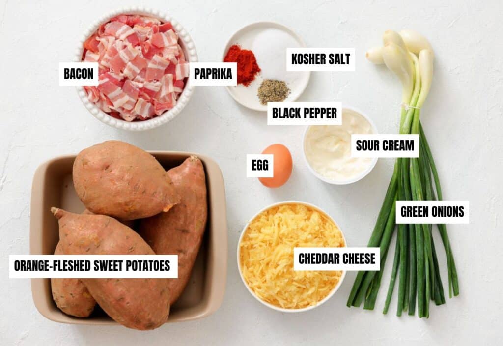 Top down view of raw ingredients for sweet potato casserole, including chopped bacon, whole sweet potatoes, shredded Cheddar, green onions, sour cream, an egg, and seasonings arranged on a white surface.