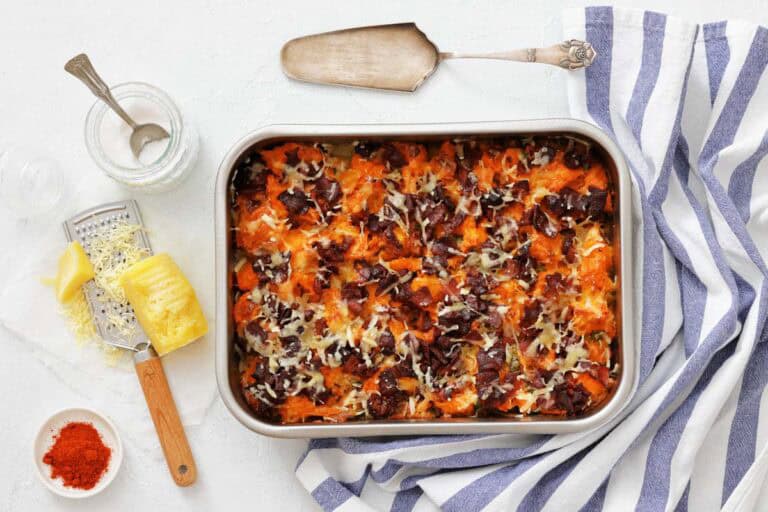 Top down view of a baked sweet potato casserole topped with crisp bacon and melted cheese in a metal pan, with grated cheese, paprika, and a serving spoon arranged around it.