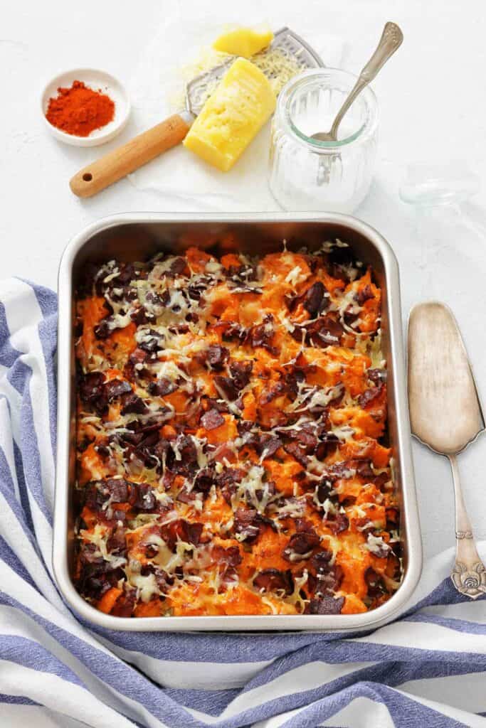A pan of baked sweet potato casserole topped with crisp bacon and melted cheese, with paprika and a block of cheese on the side.