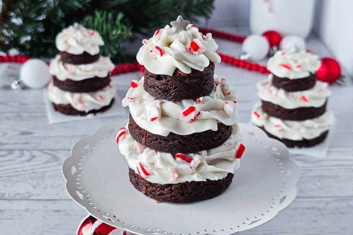 Three stacked brownie rounds with white frosting and crushed peppermint, arranged to resemble a Christmas tree, displayed on a white stand with holiday decorations in the background.