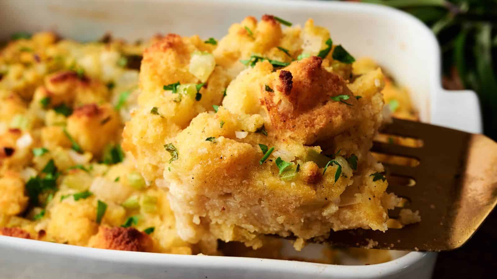A metal spatula lifts a serving of baked stuffing topped with chopped parsley from a white casserole dish.