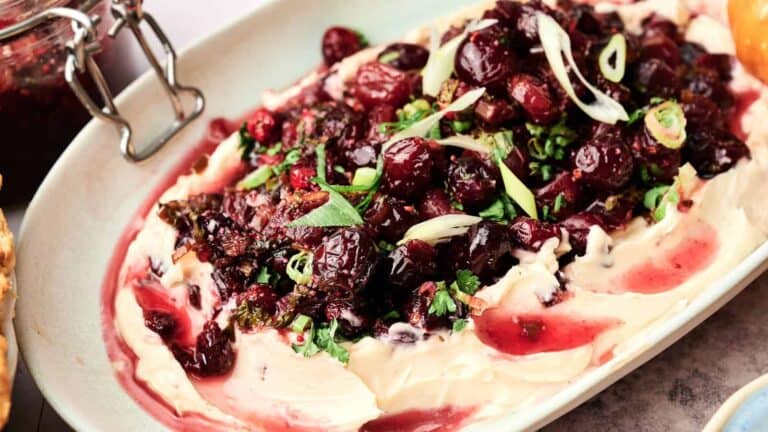 A creamy dip topped with sweetened cranberry and jalapeno sauce, garnished with fresh herbs.