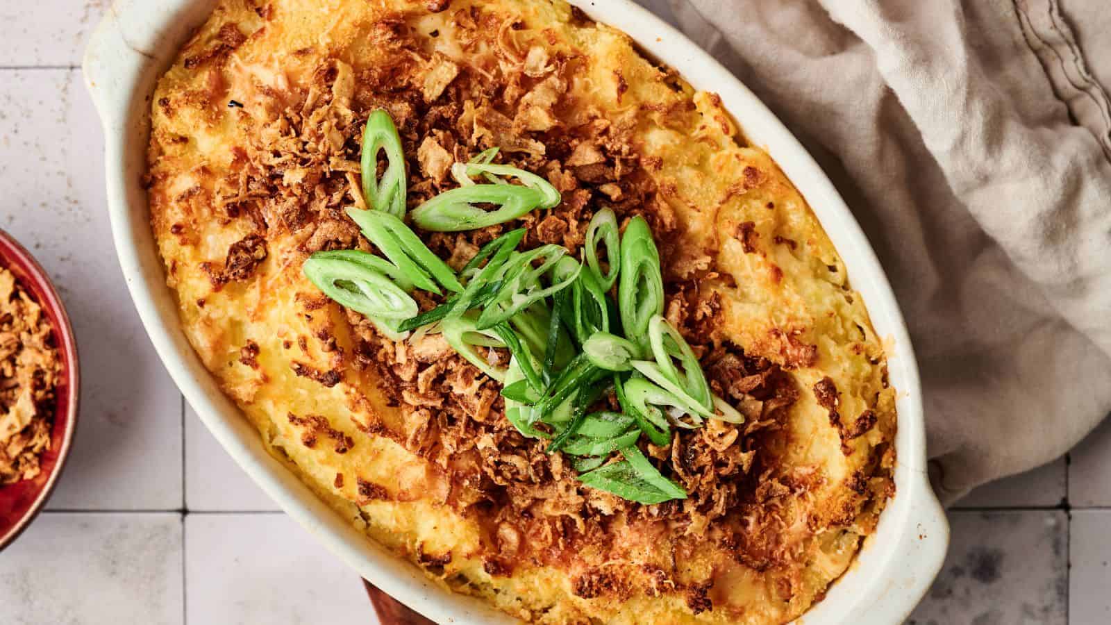 A baked casserole topped with crispy fried onions and sliced green onions, served in a white oval dish on a tiled surface with a beige cloth nearby.