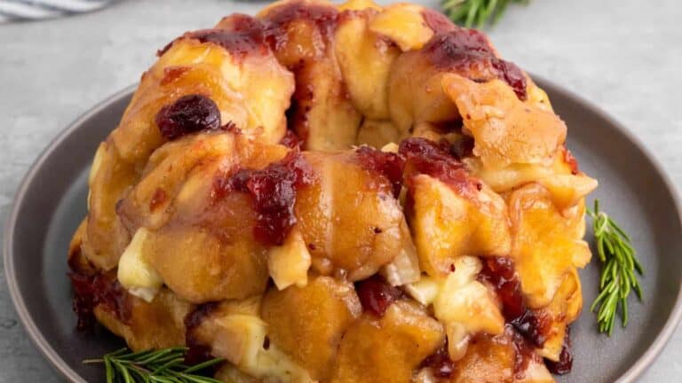 Golden-brown savory monkey bread garnished with rosemary sprigs, featuring melted cheese and cranberry sauce on a dark plate, evoking a festive holiday presentation.
