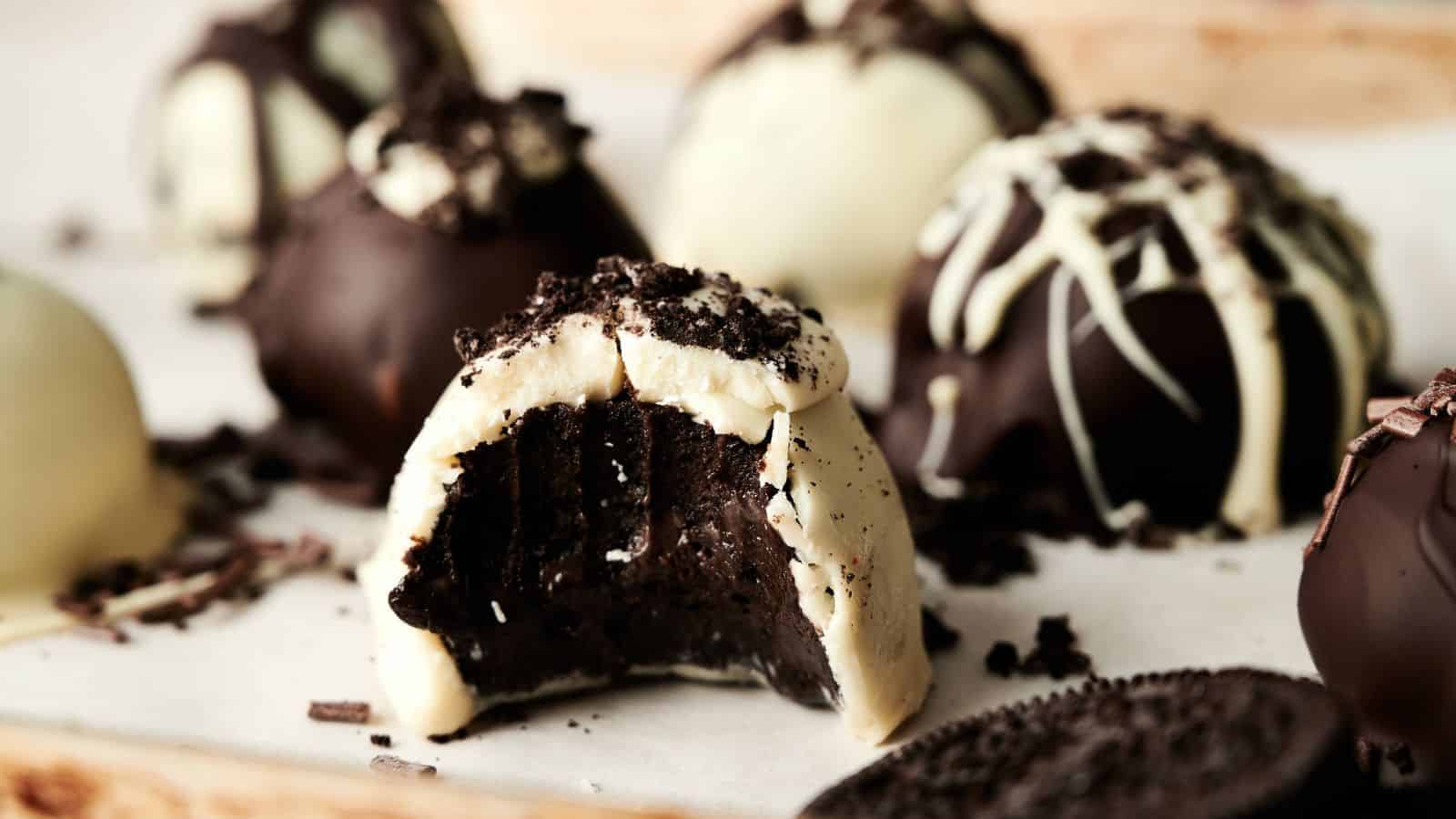 Assorted chocolate truffles, some coated in white and dark chocolate, with one bitten to show the rich chocolate filling inside. Crumbled cookies are sprinkled on top.