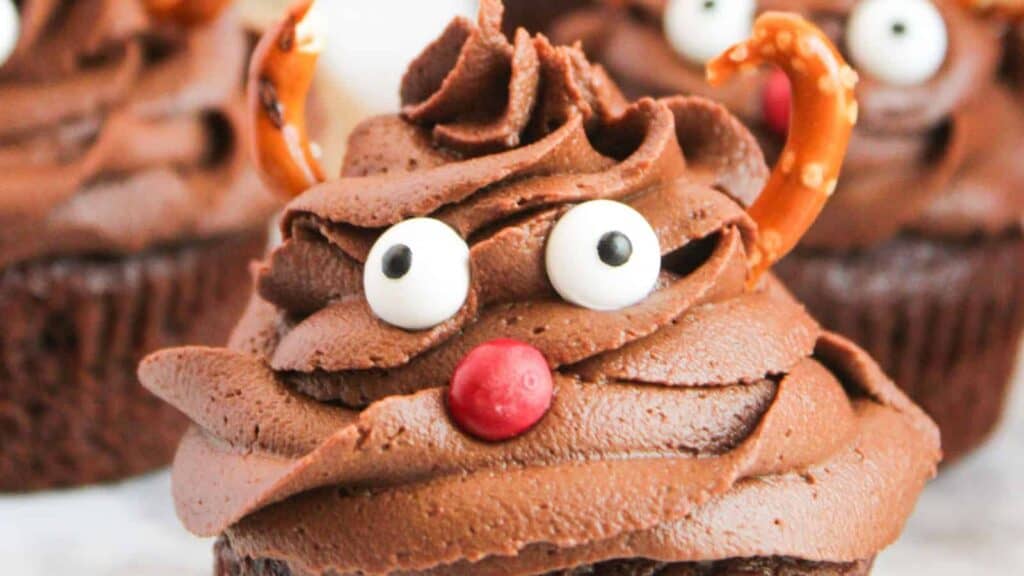 A close-up shot of reindeer-themed cupcakes with chocolate frosting, pretzel antlers, and red candy noses.