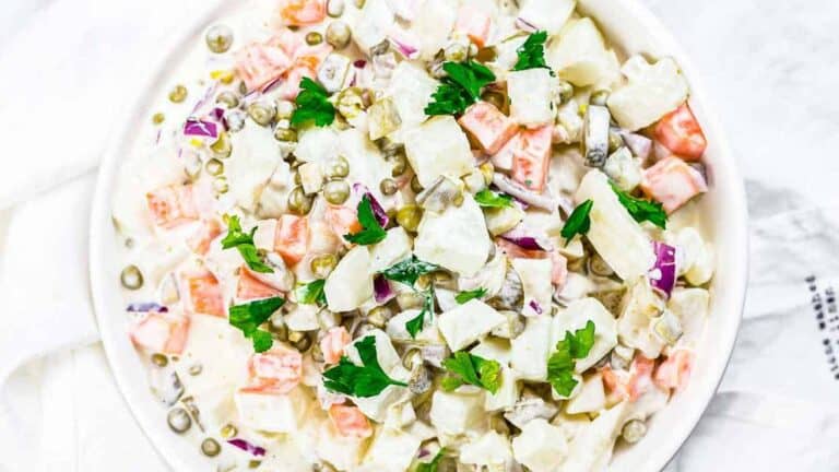 Creamy Russian salad with diced vegetables and mayonnaise, garnished with parsley in a white bowl on a white surface.