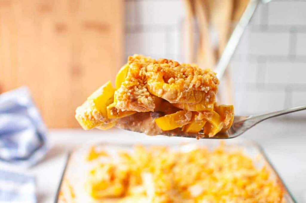 A close-up of a serving spoon holding a portion of cheesy baked casserole with yellow squash and a crumb topping, above a glass baking dish with more casserole.