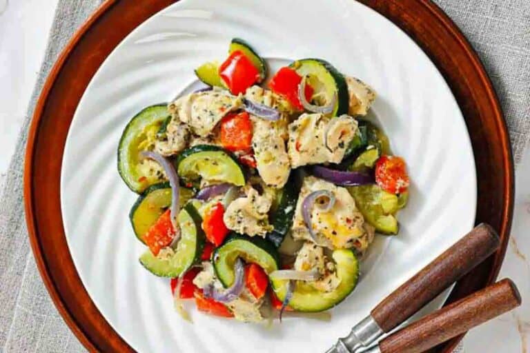 Cooked chicken pieces mixed with zucchini, red bell peppers, and red onions on a white plate with wooden utensils.