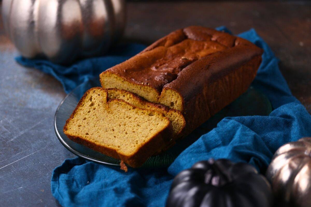 A loaf of pumpkin pound cake with two slices cut sits on a glass plate atop a blue cloth, with decorative pumpkins nearby.