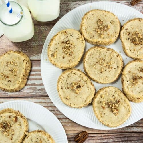 Round pecan shortbread cookies arranged on a white plate, with extra cookies, pecans, and jars of milk on a rustic wooden table.