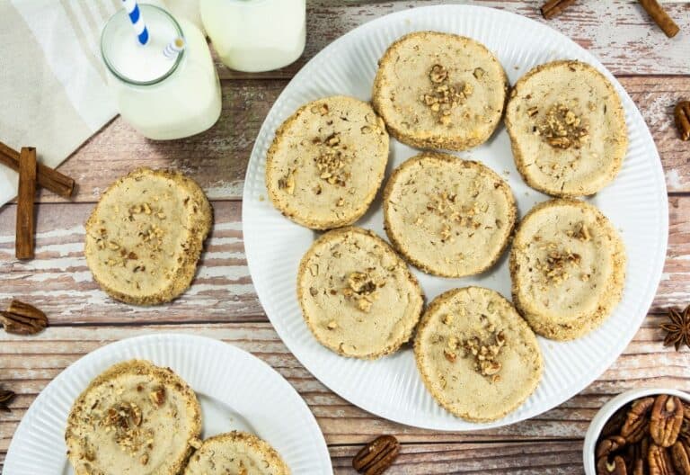 Round pecan shortbread cookies arranged on a white plate, with extra cookies, pecans, and jars of milk on a rustic wooden table.
