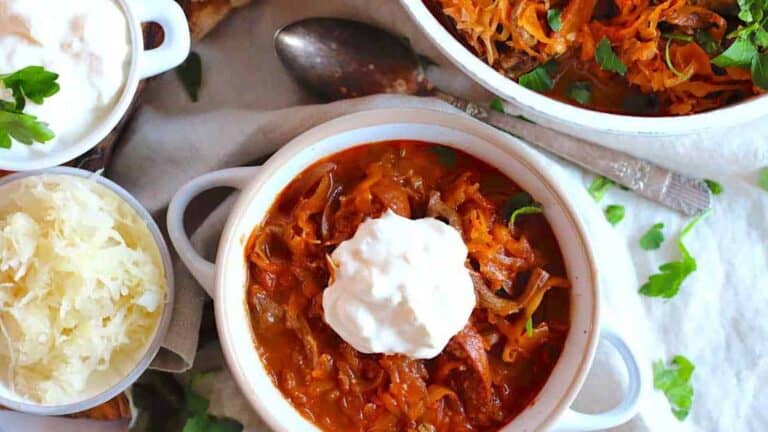 Rich red stew with shredded meat and vegetables topped with sour cream, served with sauerkraut and parsley garnish.