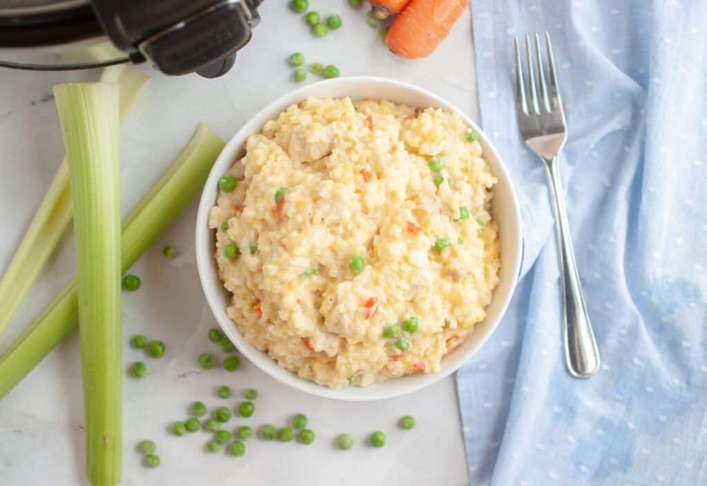 A bowl of creamy slow cooker chicken and rice with peas, surrounded by fresh celery, carrots, and scattered peas on a marble surface.