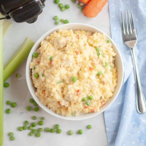 A bowl of creamy slow cooker chicken and rice with peas, surrounded by fresh celery, carrots, and scattered peas on a marble surface.