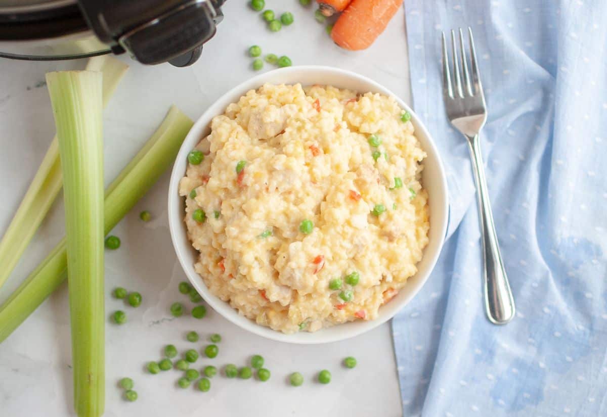 A bowl of creamy slow cooker chicken and rice with peas, surrounded by fresh celery, carrots, and scattered peas on a marble surface.