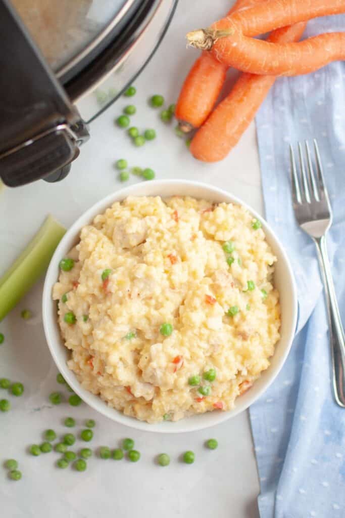 A close up bowl of creamy chicken and rice next to fresh carrots and peas, with part of a slow cooker visible in the corner.