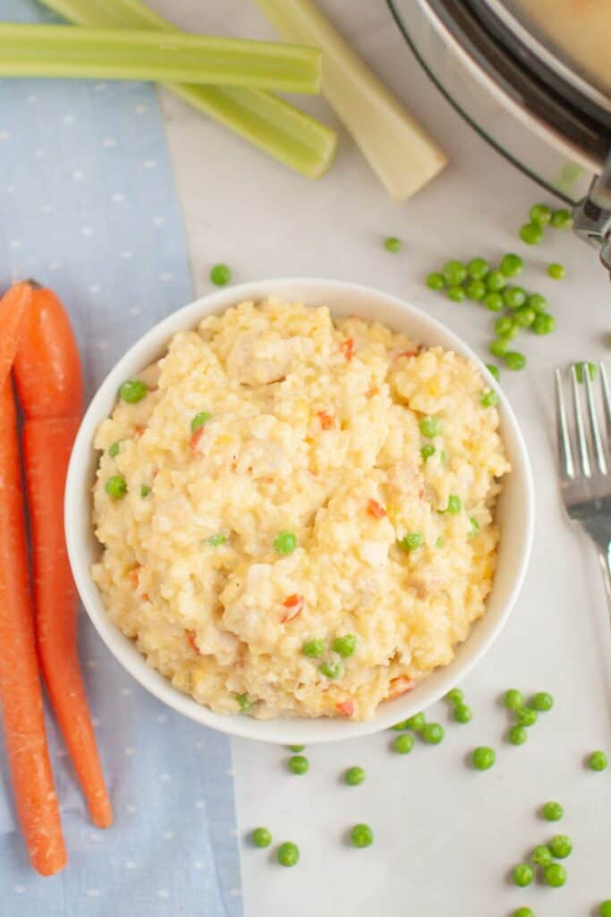 A top view of a bowl of creamy chicken and rice with peas and vegetables, surrounded by celery stalks, carrots, and scattered green peas.