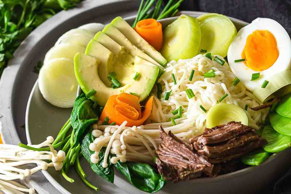 A bowl of ramen topped with sliced avocado, soft-boiled egg, shredded beef, leeks, spinach, mushrooms, and carrot slices, garnished with chopped chives.