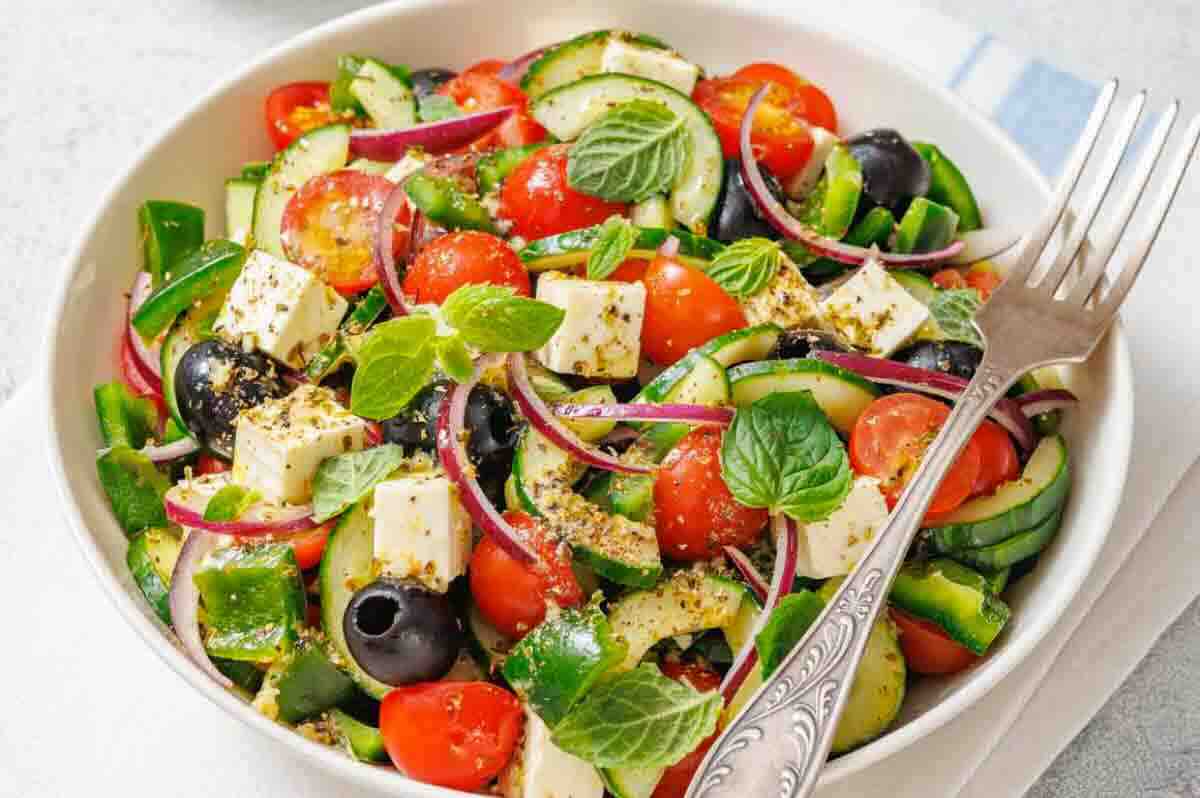 A bowl of salad with feta cheese, cherry tomatoes, cucumber, red onion, black olives, green peppers, and fresh mint, served with a fork.