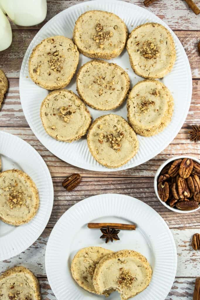 Overhead view of pecan shortbread cookies on white plates, surrounded by whole pecans, cinnamon sticks, and milk bottles on a wooden background.