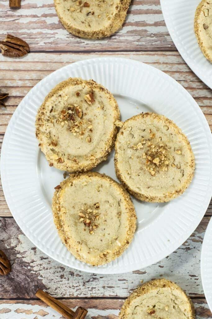 Close-up overhead shot of three pecan shortbread cookies on a white plate sitting on a weathered wooden table, with scattered pecans nearby.
