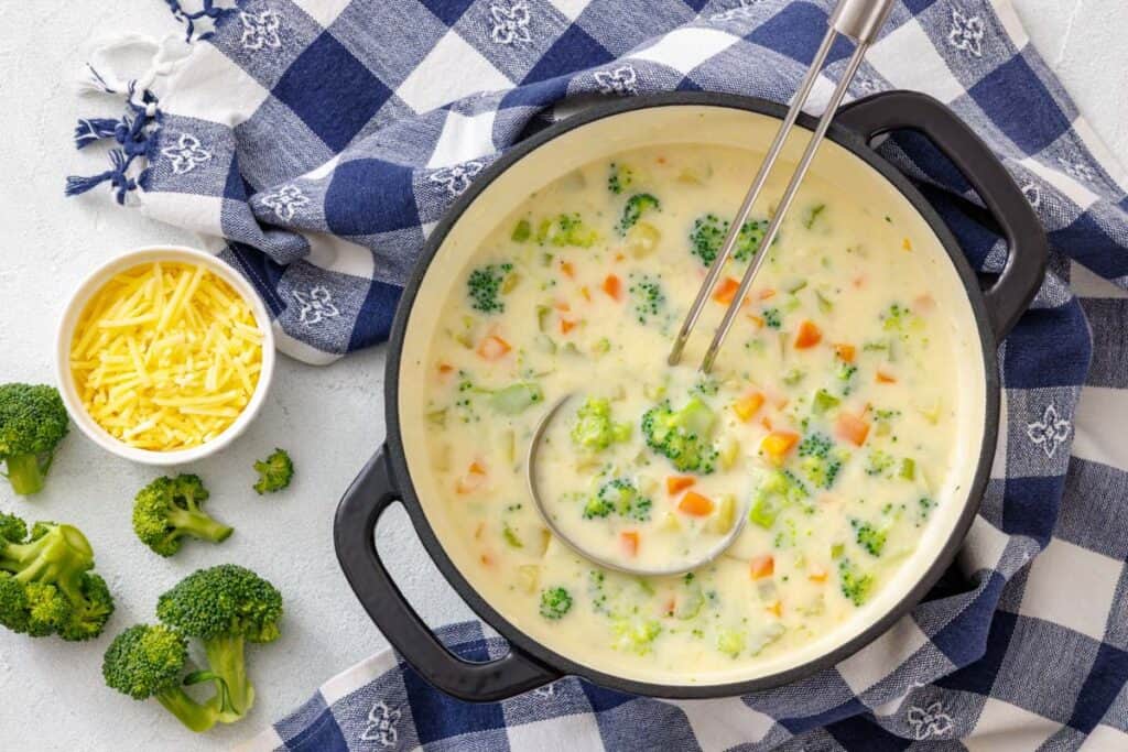 A pot of creamy vegetable chowder with broccoli, carrots, and potatoes, served with a ladle and a side of shredded cheddar cheese.