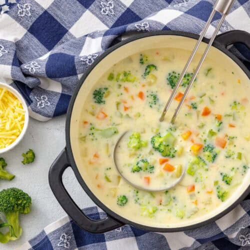 A pot of creamy vegetable chowder with broccoli, carrots, and potatoes, served with a ladle and a side of shredded cheddar cheese.