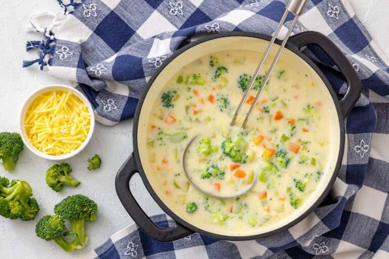 A pot of creamy vegetable chowder with broccoli, carrots, and potatoes, served with a ladle and a side of shredded cheddar cheese.