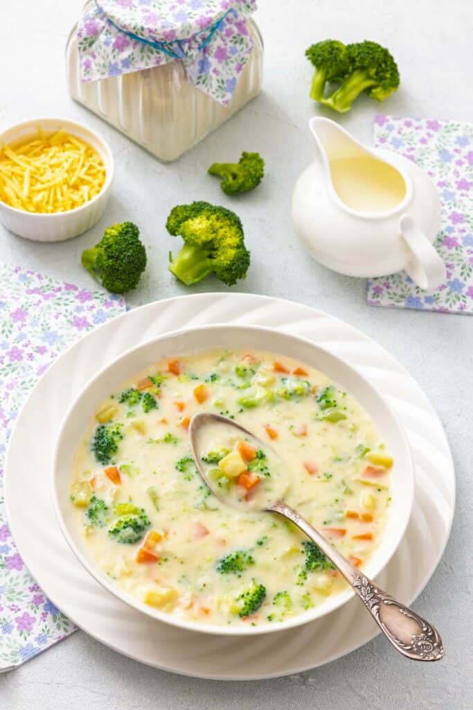 A bowl of vegetable chowder topped with fresh broccoli and carrots, served with a spoon and garnished with extra cheddar cheese.